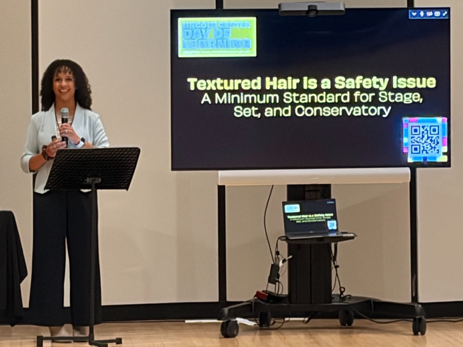 Kira Troilo smiling at a lectern with a microphone, beside a screen showing her title slide: Textured Hair Is a Safety Issue, A Minimum Standard for Stage, Set, and Conservatory.