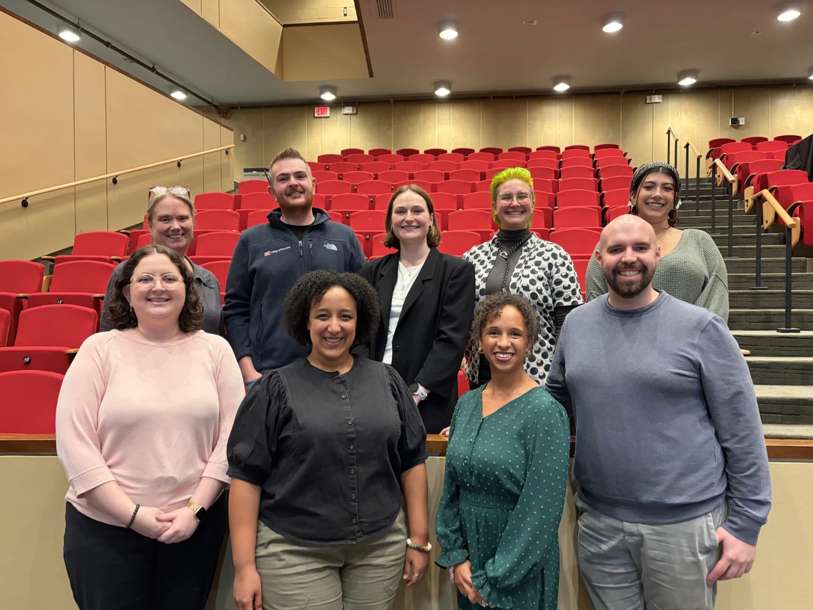 Kira Troilo with the Wheelock Family Theatre cohort, 10 people smiling together in front of the theater's red auditorium seats.